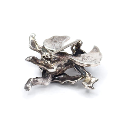 Reverse of the cherub brooch on white, showing the smooth undersides of the wings, the long pin stem crossing them, the round safety catch, and darker patina in the recesses.