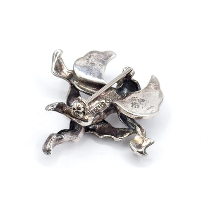 Closer crop of the reverse, tilted so the round safety catch and the pin stem lying across it sit at the centre, with the hollowed backs of the cherub's legs and feet below and patina in the folds.