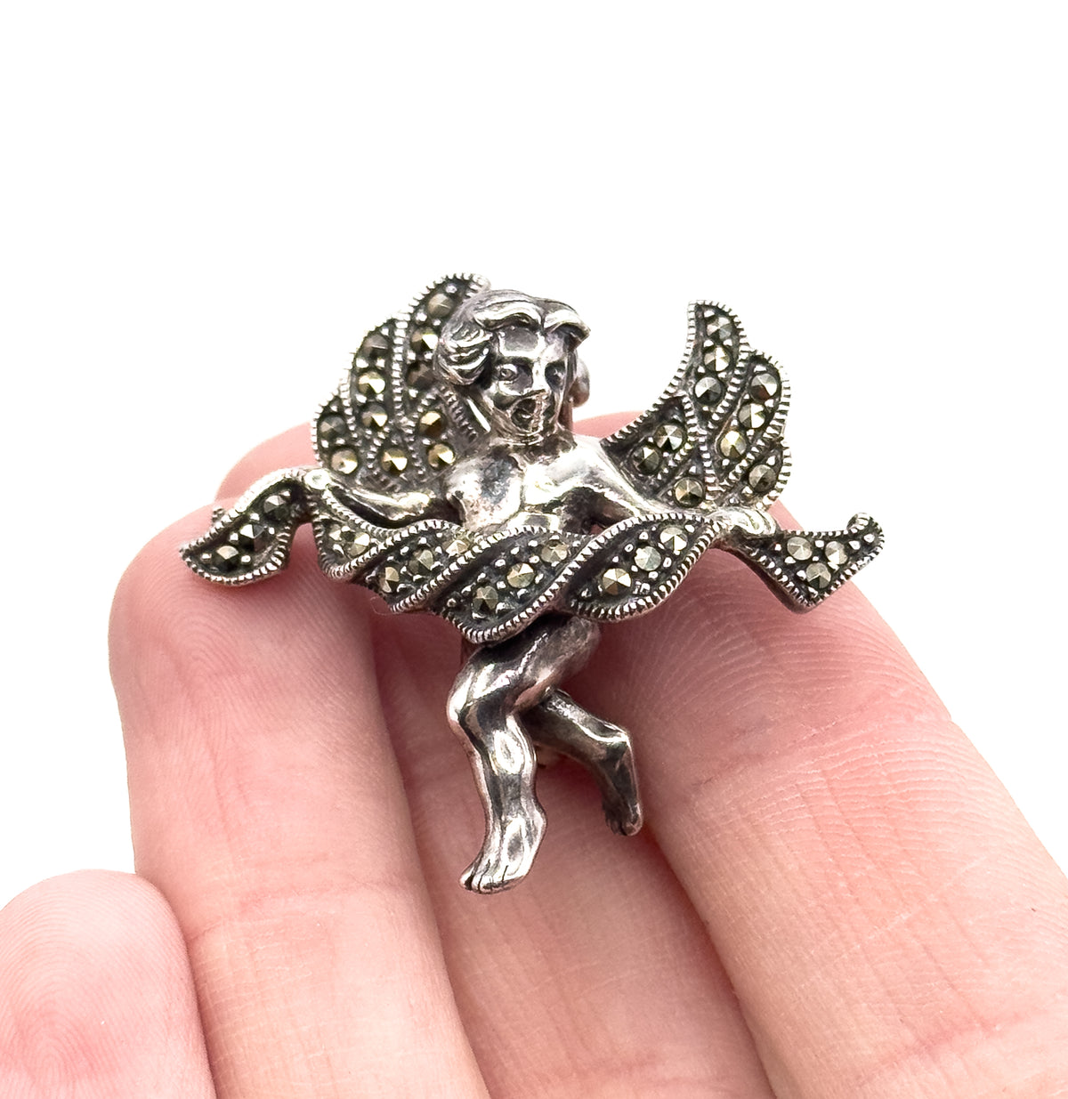 Cherub brooch resting across two fingertips, showing it spans roughly the width of two fingers, with the marcasite-set wings and draped cloth catching light from above.