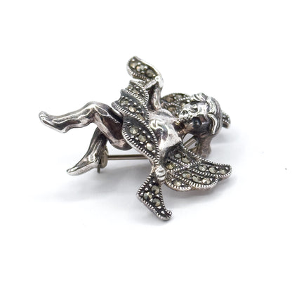 Brooch turned on its side against white so the cherub lies almost horizontal, with the straight pin stem and its catch visible behind the marcasite-set wings and draped cloth.