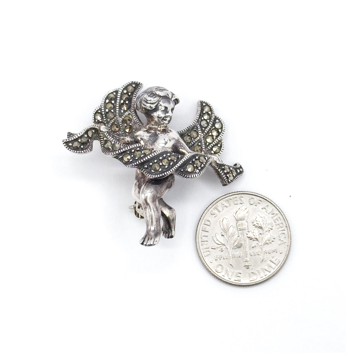 Cherub brooch laid beside a dime on white, standing a little taller than the coin's width, with the marcasite-set wings and draped cloth angled toward the camera.