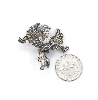 Cherub brooch laid beside a dime on white, standing a little taller than the coin's width, with the marcasite-set wings and draped cloth angled toward the camera.