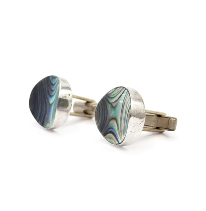 Angled three-quarter view of both faces, the concave abalone shell catching blue and green sheen beside the wide sweep of sterling silver bezel, with both toggle bars trailing to the right.