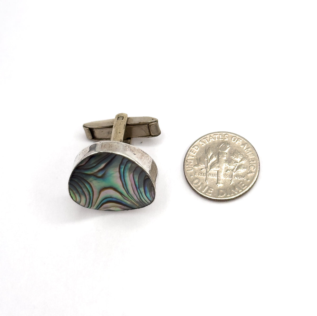 A single cufflink beside a dime for scale, its concave abalone face tilted toward the camera with the sterling silver bezel and the hinged toggle bar rising behind it.