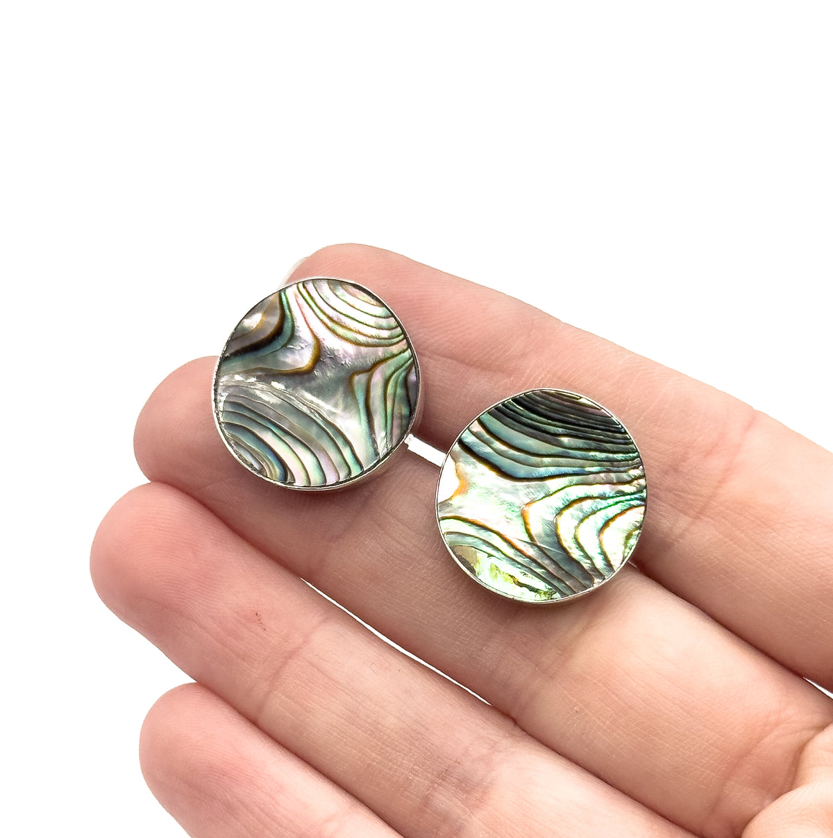 Both round faces resting side by side on open fingers, shown on the hand, each concave abalone disc in its sterling silver rim spanning roughly a fingertip for scale.
