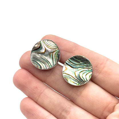 Both round faces resting side by side on open fingers, shown on the hand, each concave abalone disc in its sterling silver rim spanning roughly a fingertip for scale.