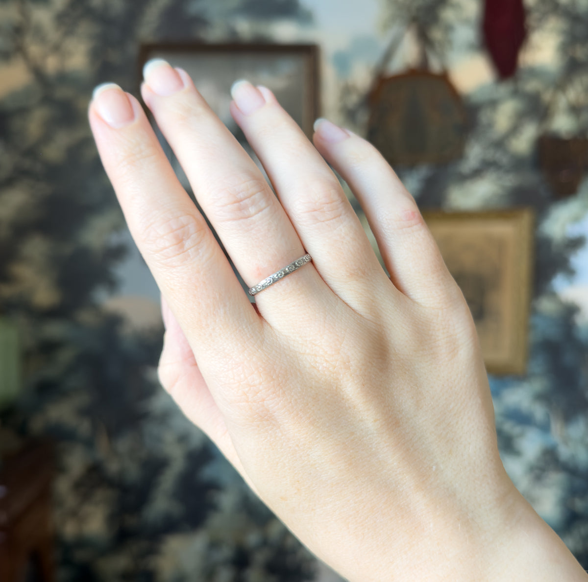 The whole hand is raised against a blurred patterned backdrop, the slim floral band on the ring finger giving a sense of its delicate scale.
