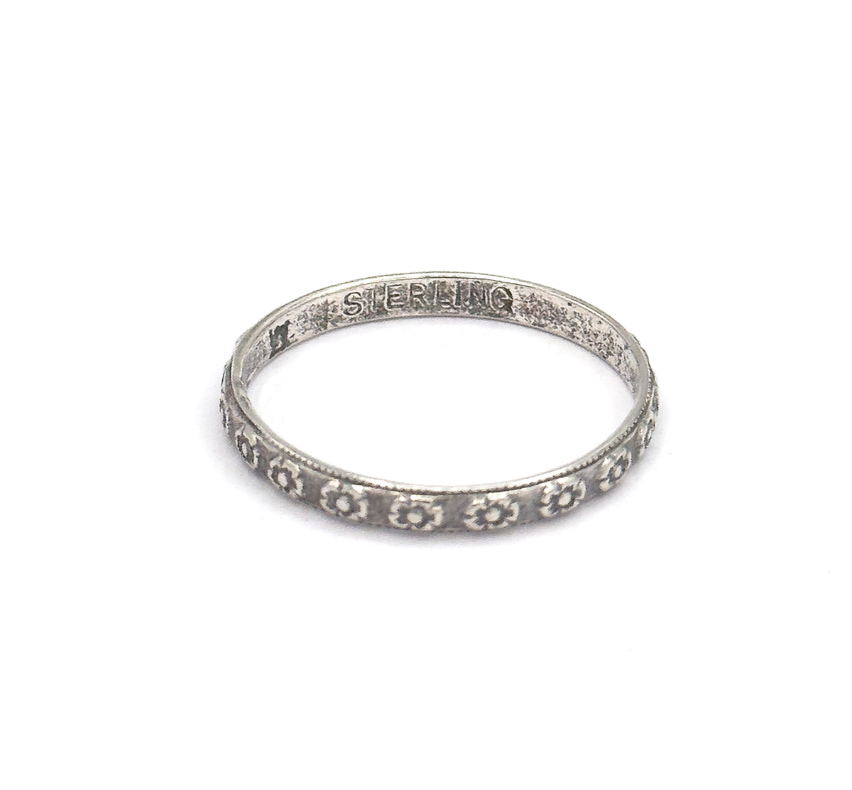 The band lies on a white background, tilted to show the flower-stamped shank in front and a stamped word inside the back of the hoop, patina darkening the recesses.