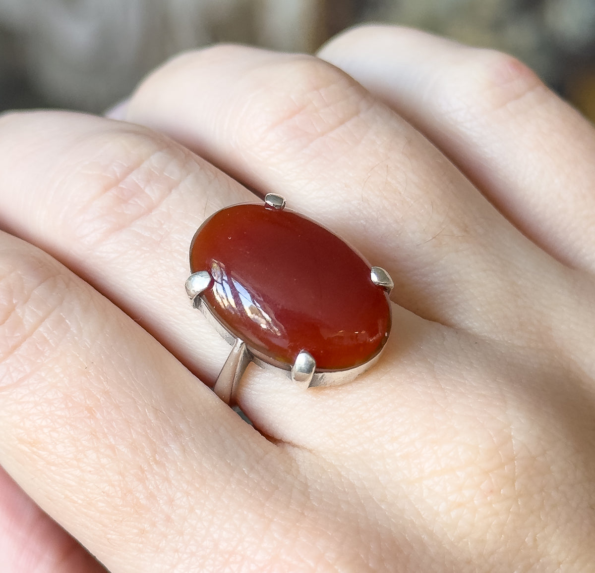 Closer view shown on the finger, the oval red cabochon filling the frame with light sliding across its domed surface and the four silver prongs gripping its edges above the narrow shank.