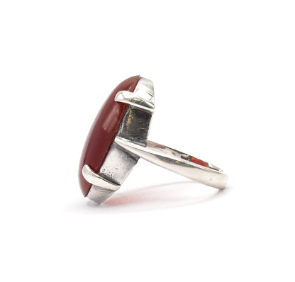Edge-on from the opposite side, the flat silver bezel wall and two prongs grip the red cabochon's rim while the shank sweeps back to the right.