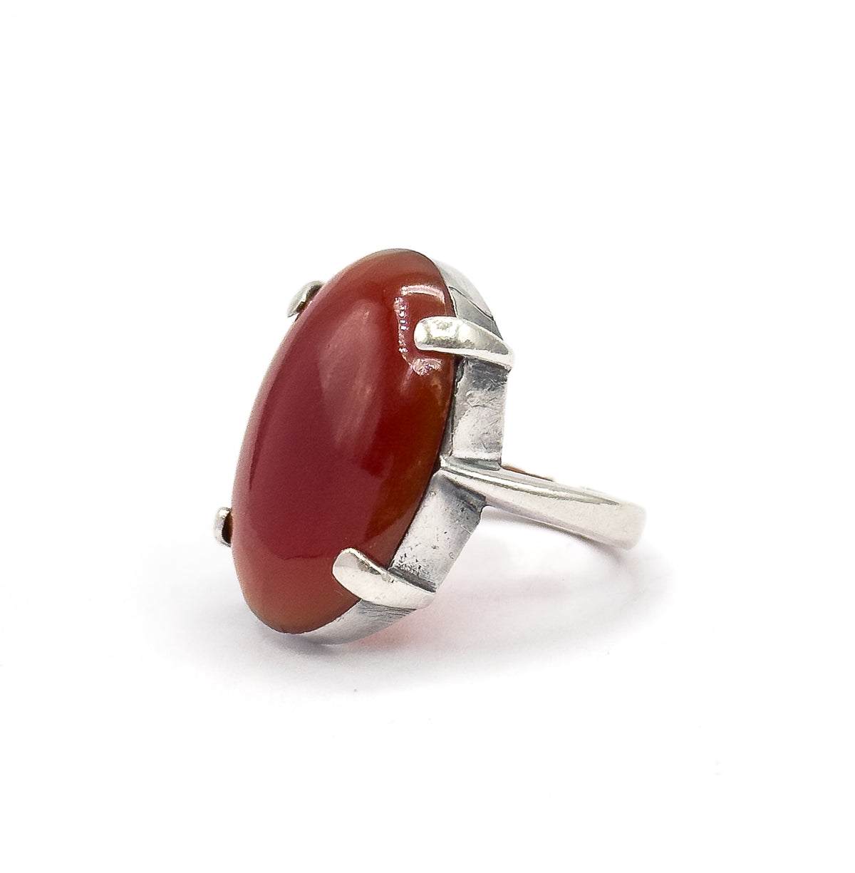 Three-quarter angle with the oval red cabochon tilted toward the viewer, its silver prongs reaching across top and bottom of the stone and the shank receding to the right.