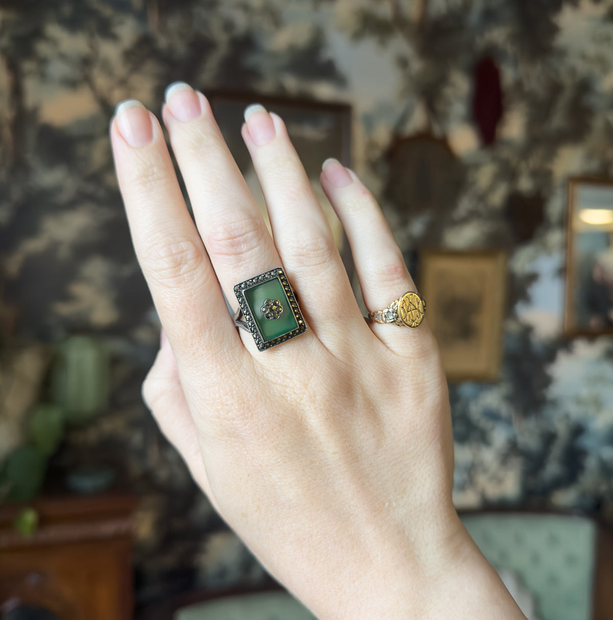The ring shown on the middle finger of a raised hand against a soft furnished background, its green rectangular face and marcasite halo sitting clearly proud of the knuckle.
