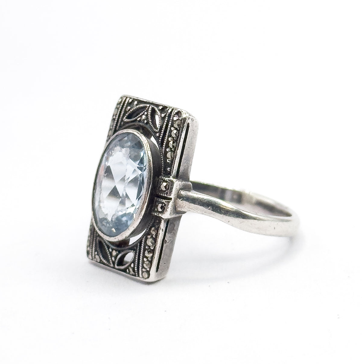 Angled three-quarter view from the front left, the oval icy blue stone and its bezel tilted toward the camera while the marcasite-set border and shoulders recede to the right.