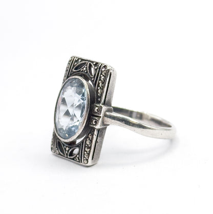 Angled three-quarter view from the front left, the oval icy blue stone and its bezel tilted toward the camera while the marcasite-set border and shoulders recede to the right.