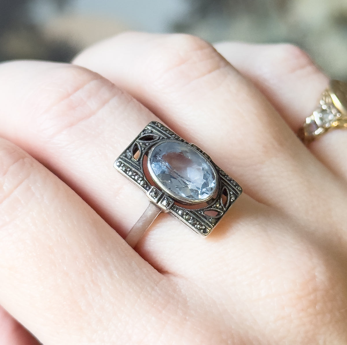 Sterling silver ring with an oval icy blue synthetic spinel bezel-set in a marcasite-studded openwork rectangular face, shown on a finger to convey its size.