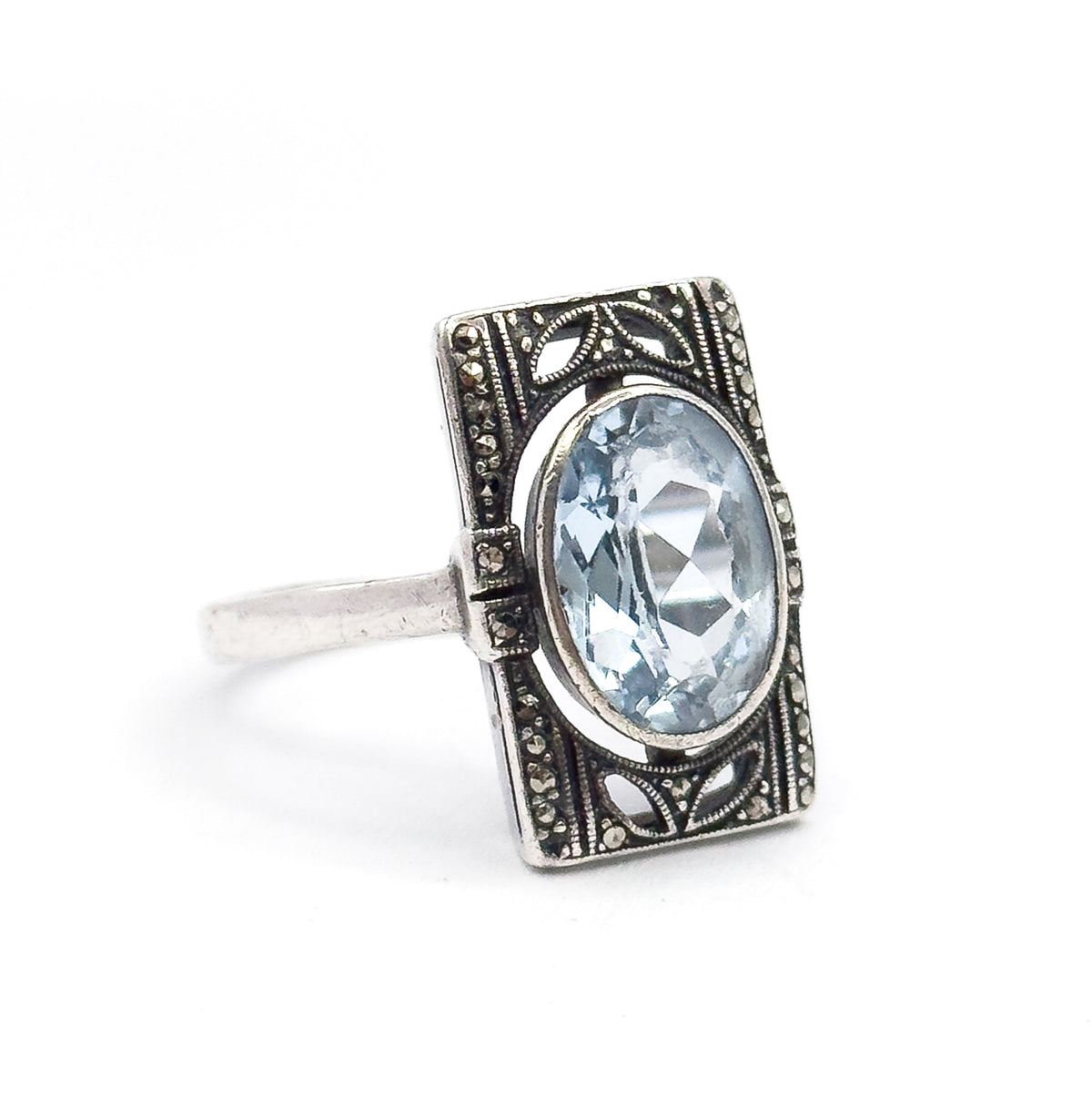 Off the hand against white, the ring is angled so the oval icy blue stone and its bezel face the viewer while the slim sterling shank curves away behind the marcasite-set rectangular frame.