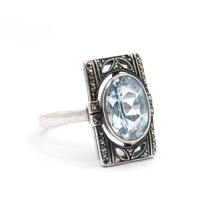 Off the hand against white, the ring is angled so the oval icy blue stone and its bezel face the viewer while the slim sterling shank curves away behind the marcasite-set rectangular frame.