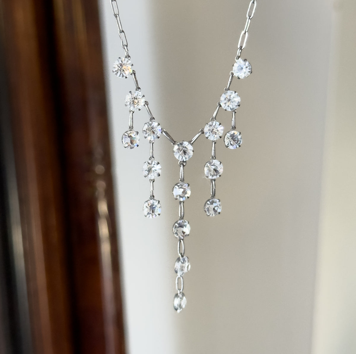 Hung in mid-air against a softly blurred pale wall beside a dark wooden frame edge, the fringe of paste drops swinging free so the central tapering strand hangs longest.