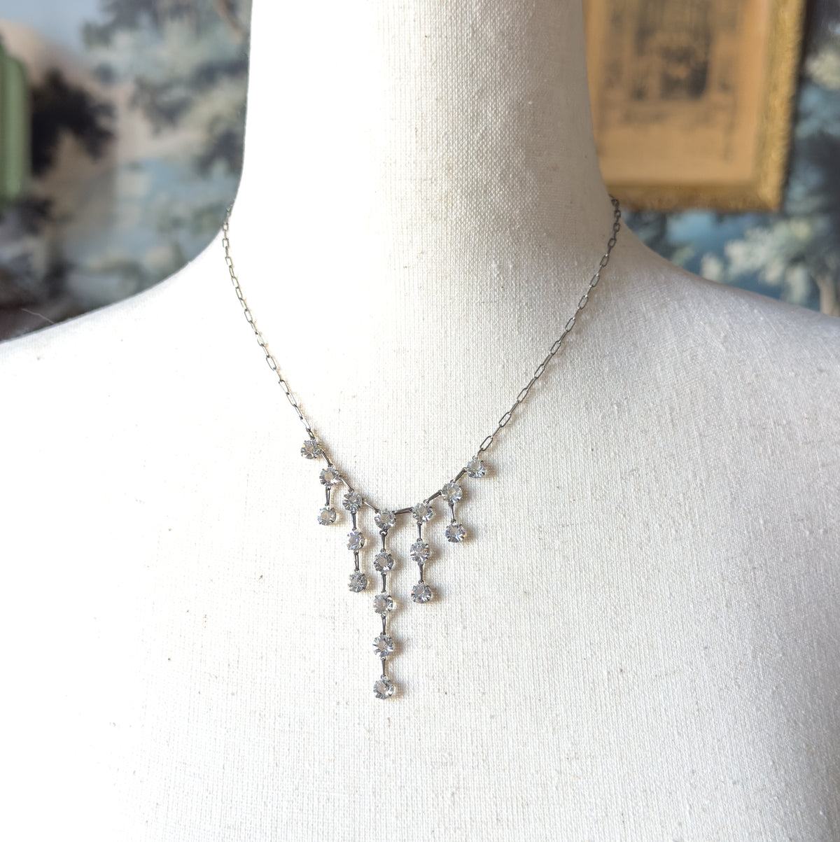 Art Deco paste festoon fringe necklace shown at the neck on a pale cloth bust form, its fine chain meeting graduated drops of bright paste stones that fall in a tapering fringe.