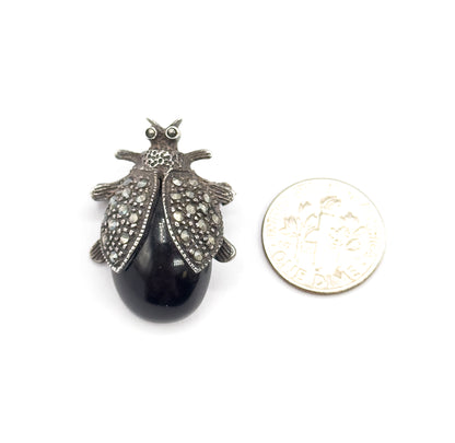 Top view beside a small coin for scale, the brooch only a little larger than the coin, its black cabochon body and small-stone-set wings shown against white.