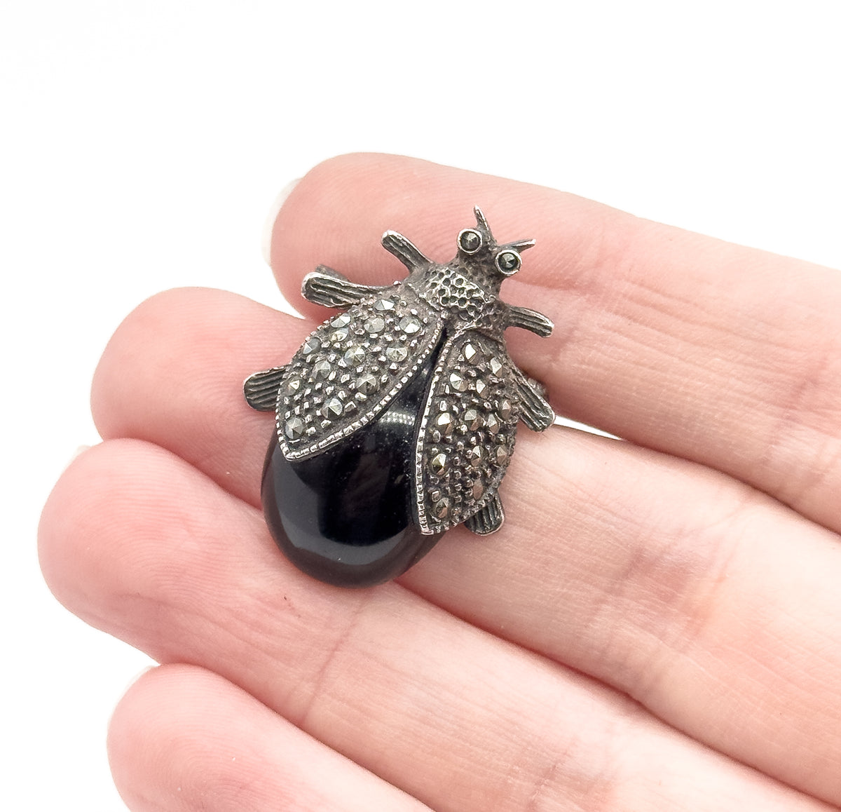 Vintage sterling silver jelly belly bug brooch, its glossy black cabochon body framed by sparkling-stone-set wings, shown resting across the fingers of a hand for scale.