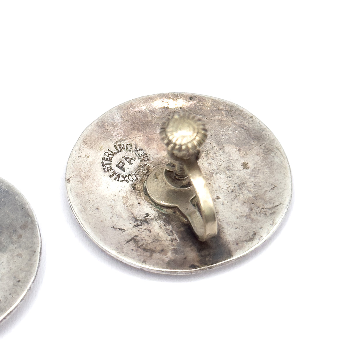 Reverse of one disc fills the frame, showing the screwback fitting with its ridged knob and curved arm beside an impressed circular maker's stamp, the second earring at the edge.