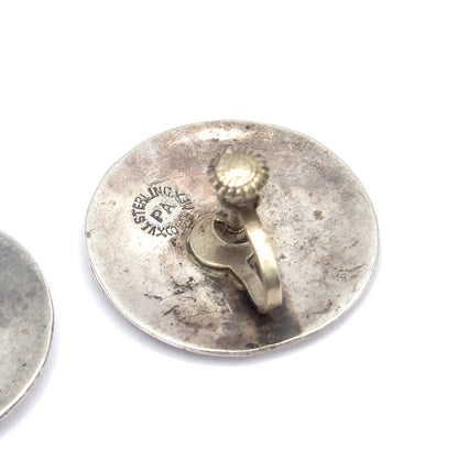 Reverse of one disc fills the frame, showing the screwback fitting with its ridged knob and curved arm beside an impressed circular maker's stamp, the second earring at the edge.