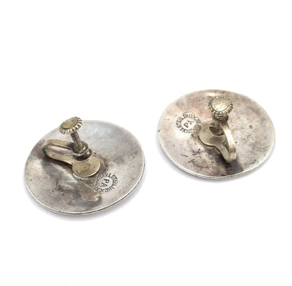 Both discs seen from behind at an angle, each curved sterling back carrying a ridged screwback knob, threaded post and flat paddle arm, with circular stamps on both.