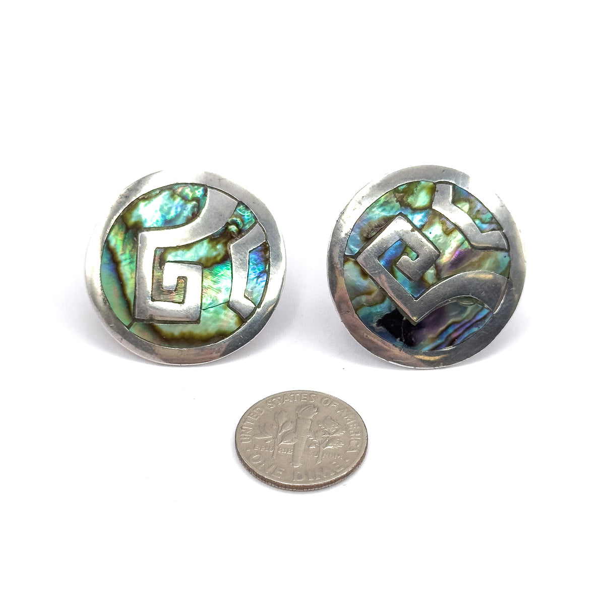 Both discs face up side by side above a dime, the coin showing how much wider each abalone face is, with angular silver scrollwork crossing the shimmering shell.