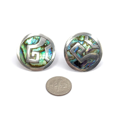 Both discs face up side by side above a dime, the coin showing how much wider each abalone face is, with angular silver scrollwork crossing the shimmering shell.