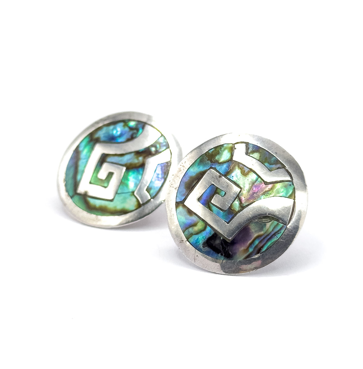 Both earrings angled upright and overlapping at a low viewpoint, so the silver disc rims stand proud as raised bezels around abalone glowing green, blue and purple.