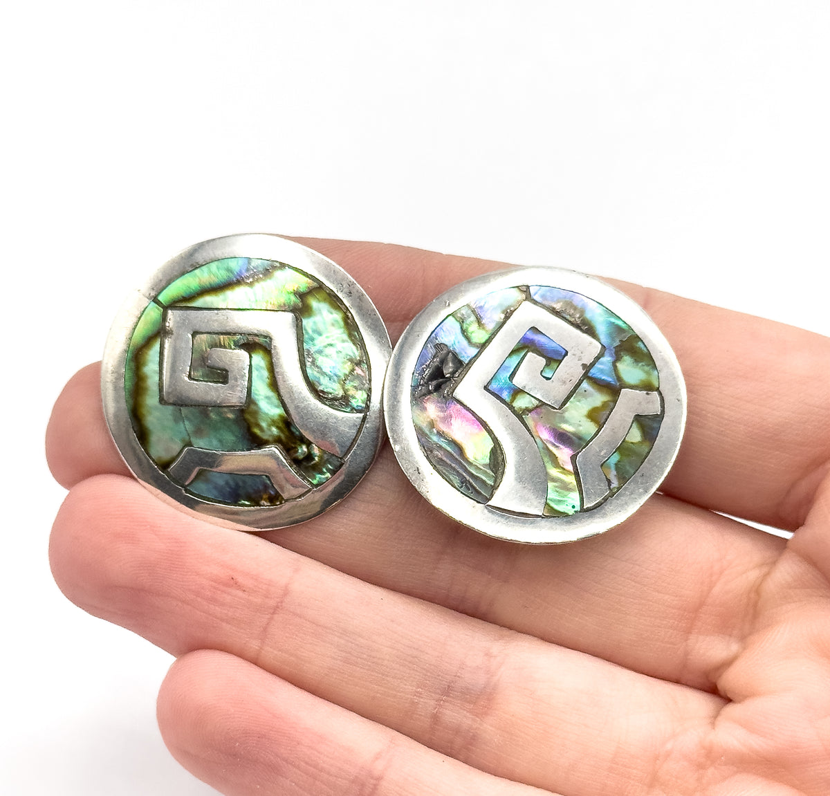 Mid-century abalone shell disc earrings in sterling silver, the pair shown resting on open fingers which convey their size, each round face set with stepped scrolling silver motifs over iridescent shell.