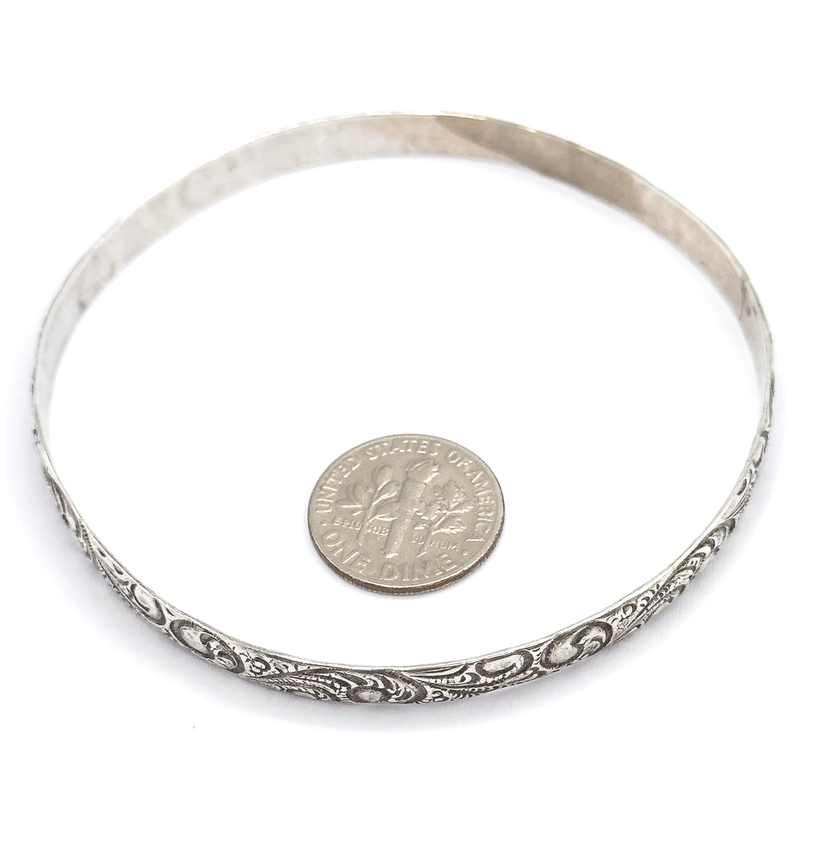 The bangle lies flat, viewed from above with a dime inside the circle for scale, the raised scrollwork and patina running along the front edge of the band.