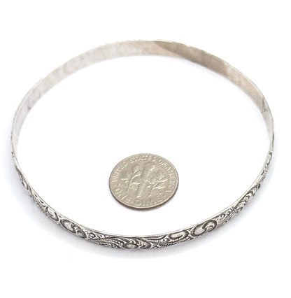 The bangle lies flat, viewed from above with a dime inside the circle for scale, the raised scrollwork and patina running along the front edge of the band.