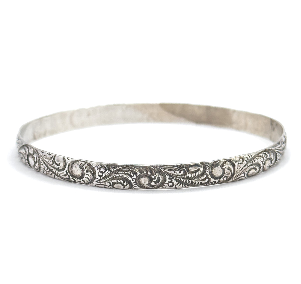 Side-on view at eye level, the bangle standing as a narrow oval with the scrollwork running sharply in focus across the front and the plain back of the band curving behind.