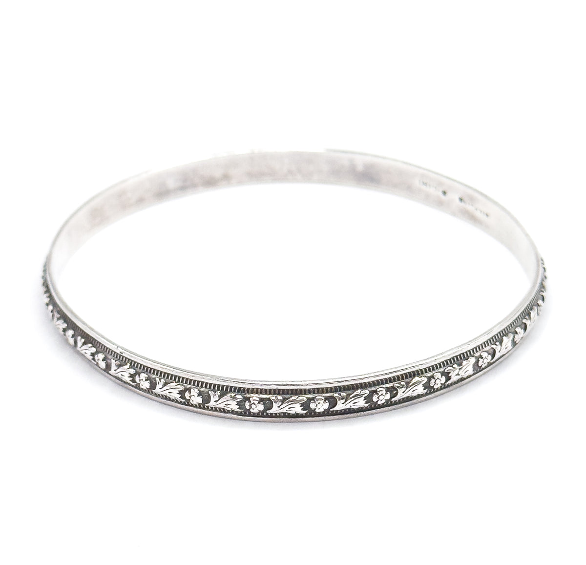 The bangle tilted lower so the carved flower-and-leaf station and its beaded borders run across the front rim, with the smooth inner wall and faint stamp visible at the back.