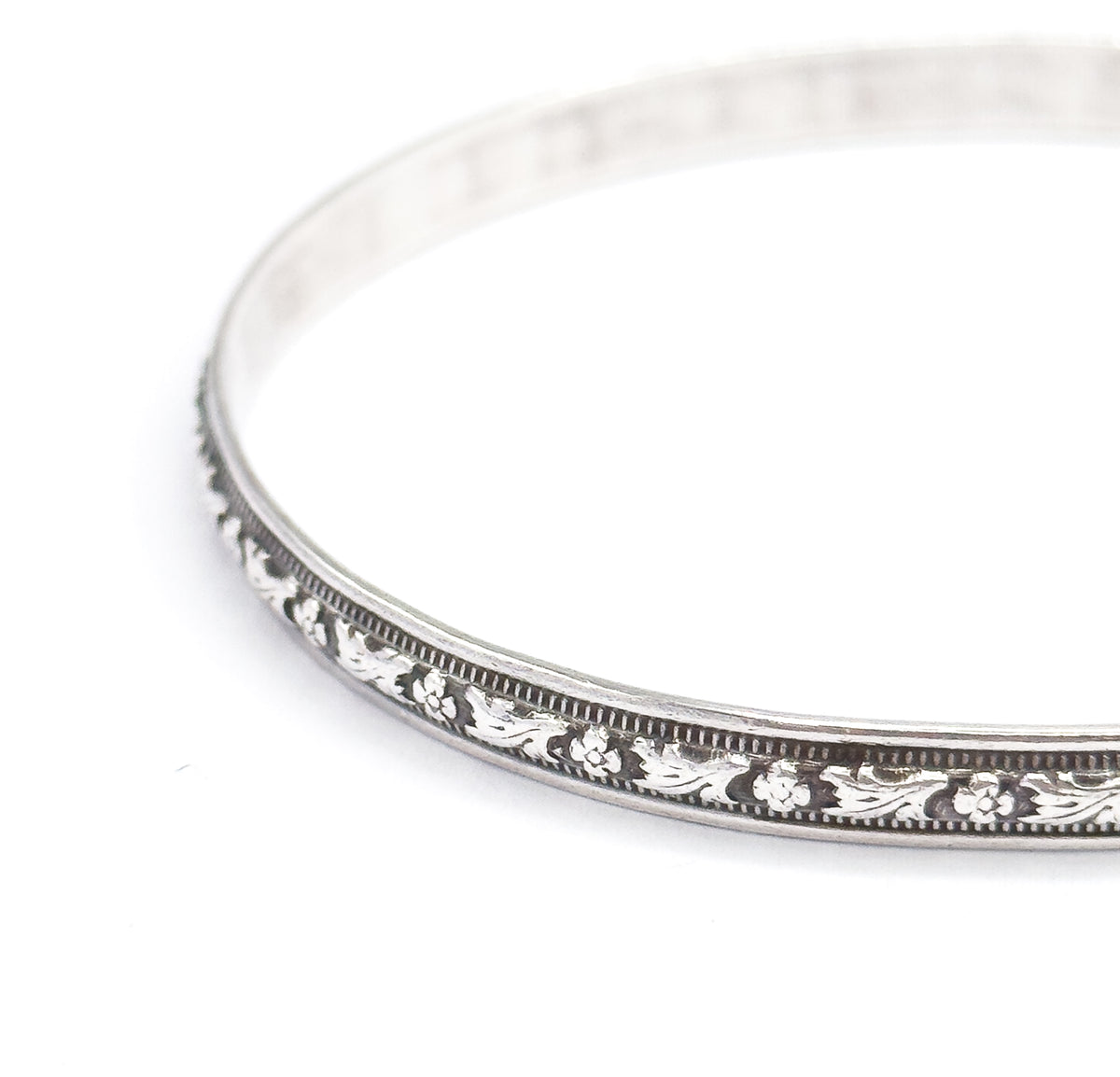 Low three-quarter view of the whole bangle tipped away from the camera, showing how slim the band is edge-on as the carved flower station and beaded borders curve off to the right.