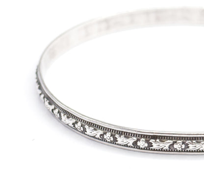Low three-quarter view of the whole bangle tipped away from the camera, showing how slim the band is edge-on as the carved flower station and beaded borders curve off to the right.