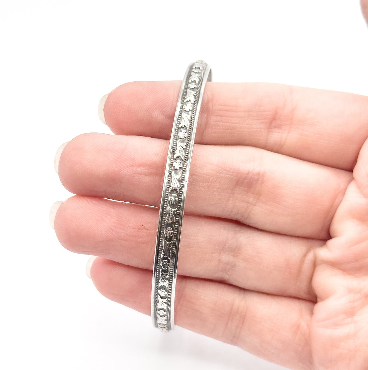 A sterling silver floral bangle shown resting across the fingers of an open hand, its narrow band edge-on so the carved flower-and-leaf design and beaded borders face the viewer.
