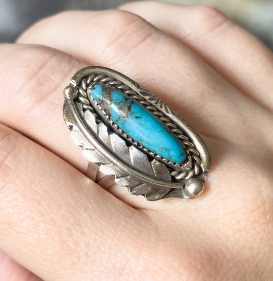 Shown on a finger, the ring's long oval turquoise cabochon and feathered sterling silver frame span nearly the full width of the finger, with rounded silver beads at the shoulders.
