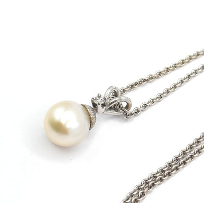 Angled view from slightly above with the chain doubling back on itself in two strands across the lower right, the ribbon-loop bail and its small diamond turned toward the pearl below.
