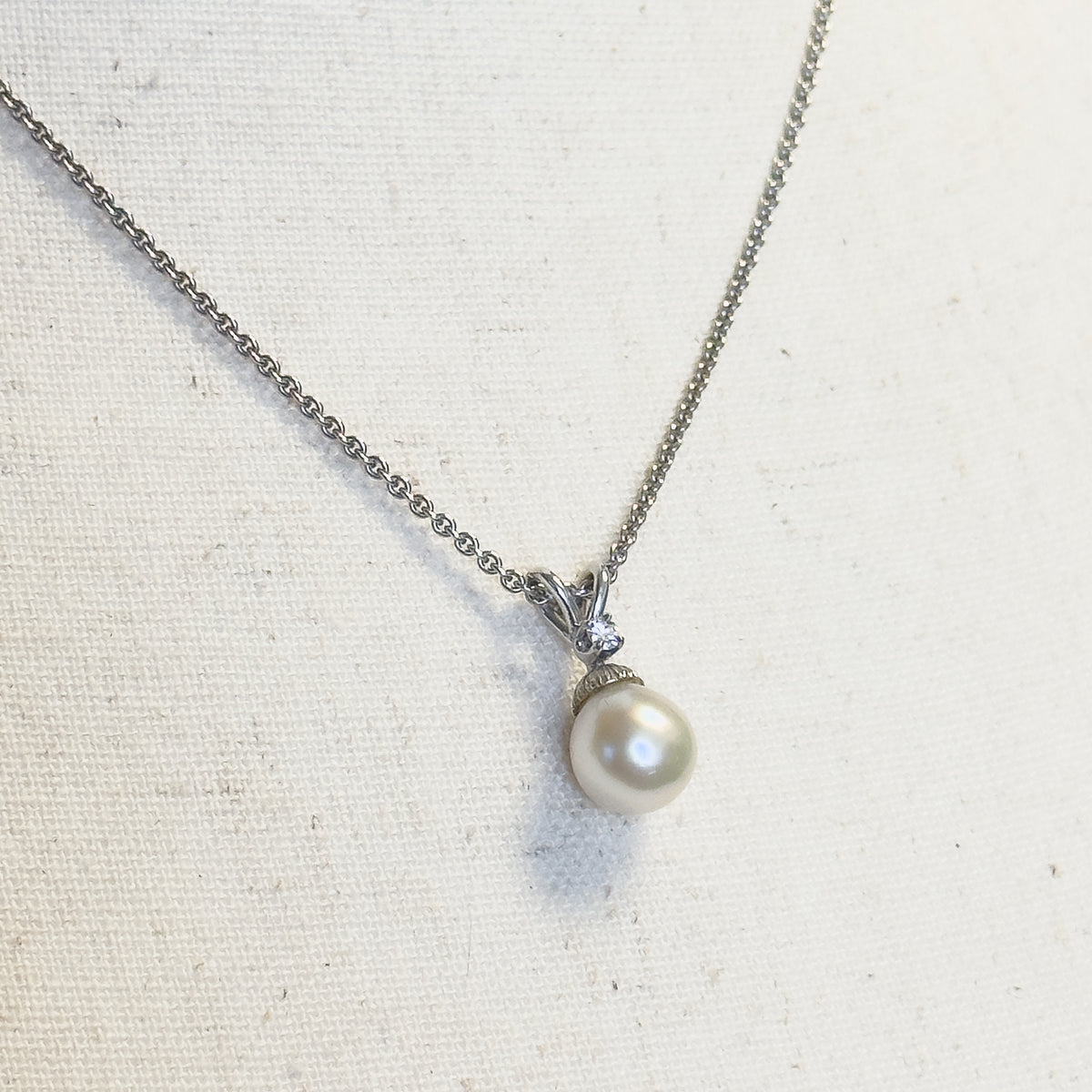 A cultured freshwater pearl and diamond pendant necklace in white gold, shown laid flat on cream linen with the fine chain curving up from the pearl and its diamond-set bail.