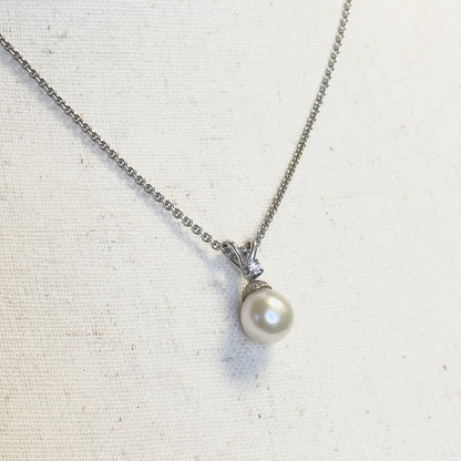 A cultured freshwater pearl and diamond pendant necklace in white gold, shown laid flat on cream linen with the fine chain curving up from the pearl and its diamond-set bail.
