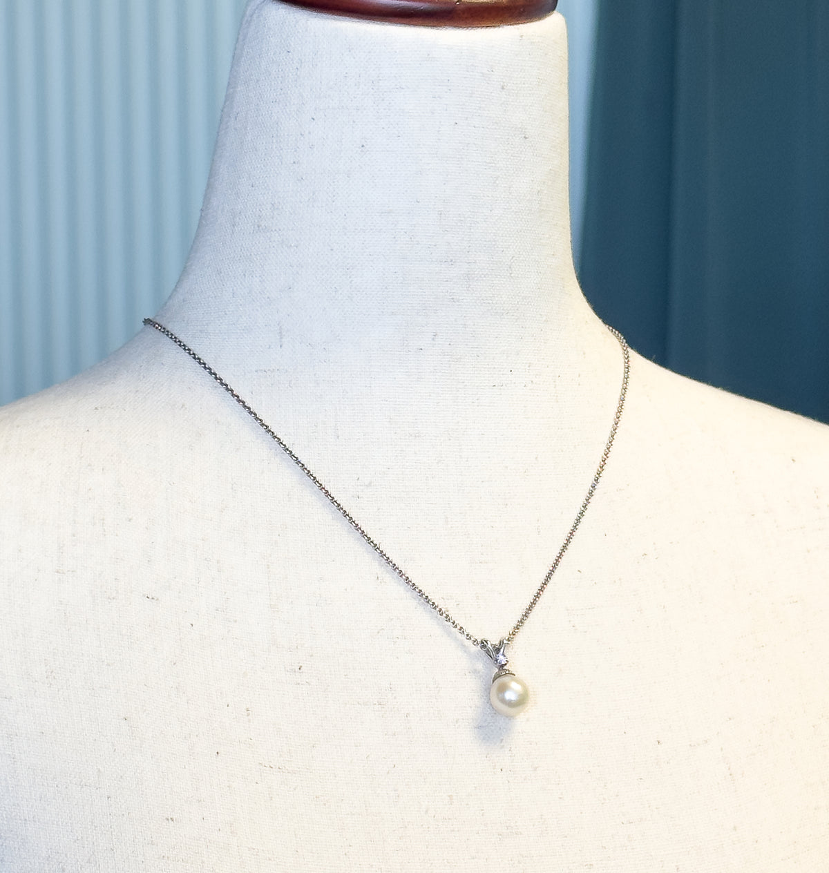 Shown at the neck on a cream linen display form, the fine chain tracing the collarbones and the creamy pearl with its diamond-set bail resting just below the hollow of the throat.