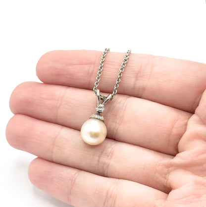 The pendant shown on the fingers of an open hand for scale, the creamy pearl spanning roughly half a fingertip with the diamond bail and chain above it.