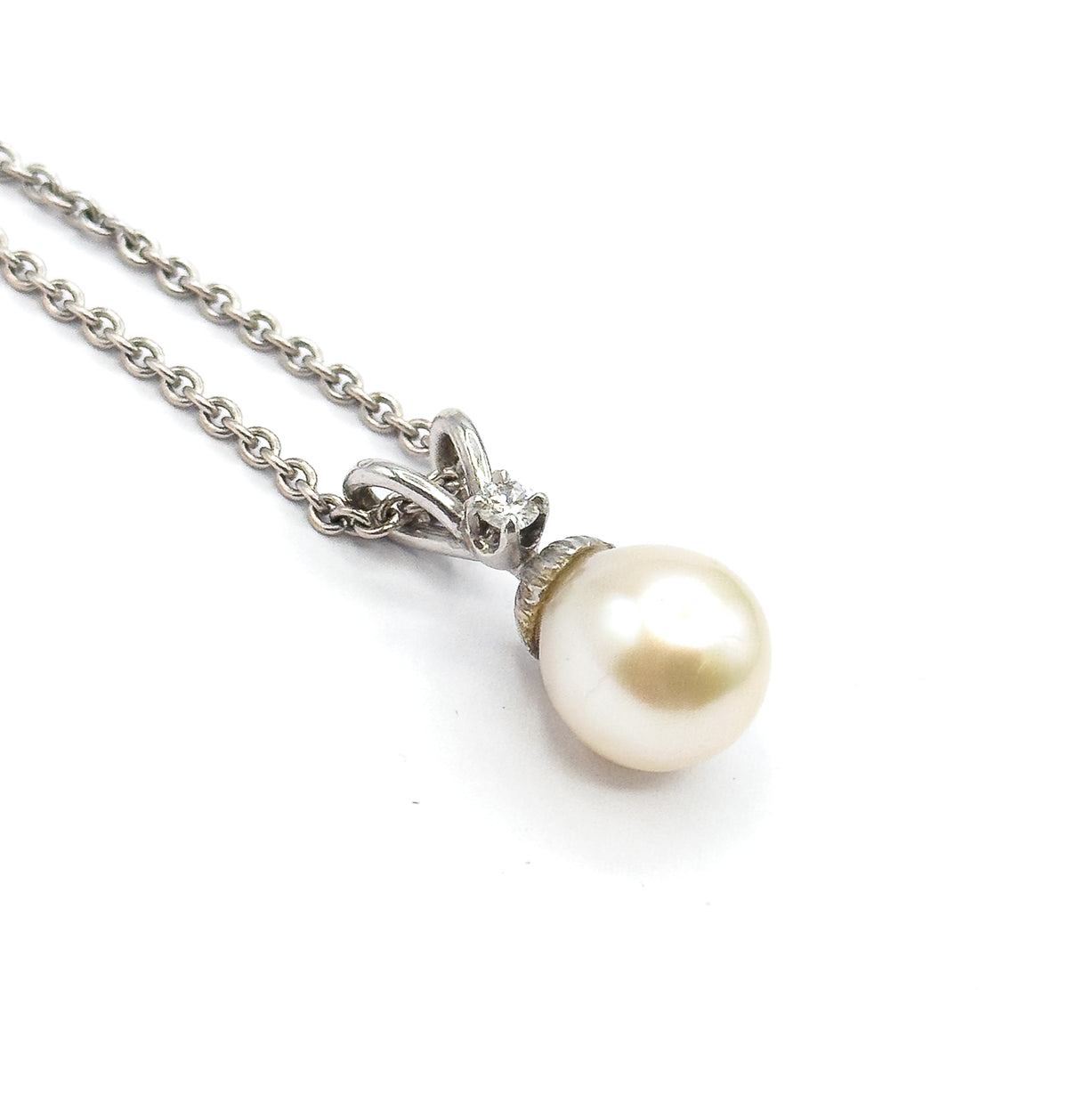 Angled close view against white showing the diamond glinting in its four-prong setting on the ribbon-like bail, with the creamy pearl below and the chain trailing off to the upper left.