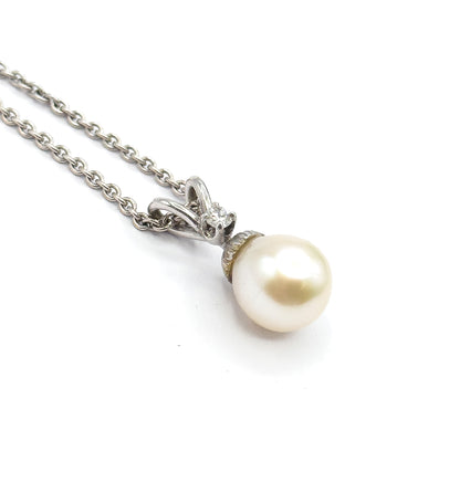 Angled close view against white showing the diamond glinting in its four-prong setting on the ribbon-like bail, with the creamy pearl below and the chain trailing off to the upper left.
