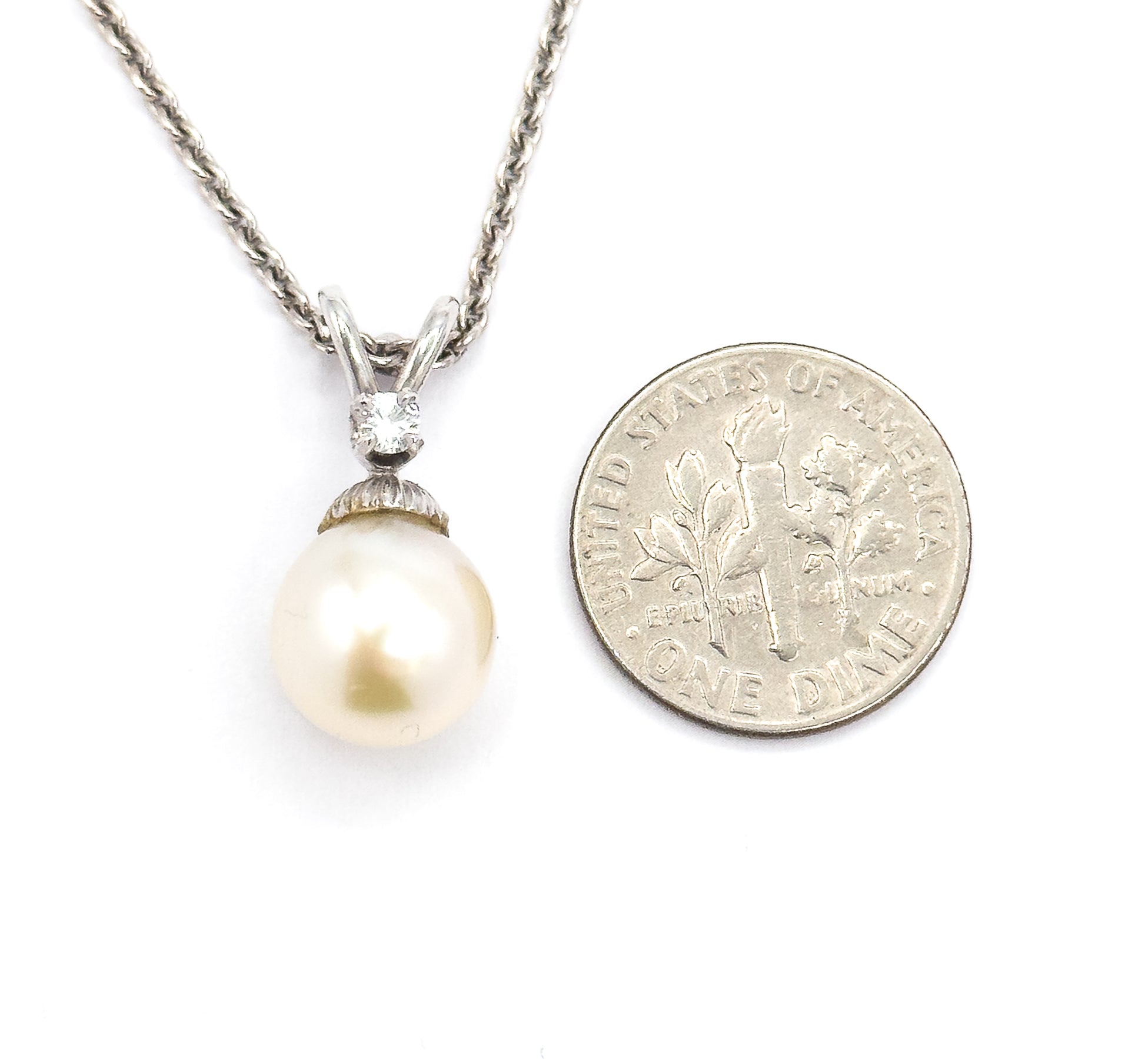 The pendant laid beside a dime for scale, the creamy pearl and diamond bail together standing about as tall as the coin is wide, chain curving up to the left.