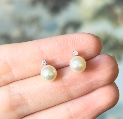 Both earrings rest on fingertips, giving a sense of their small size, each creamy pearl facing forward with a prong-set diamond above it in white gold.