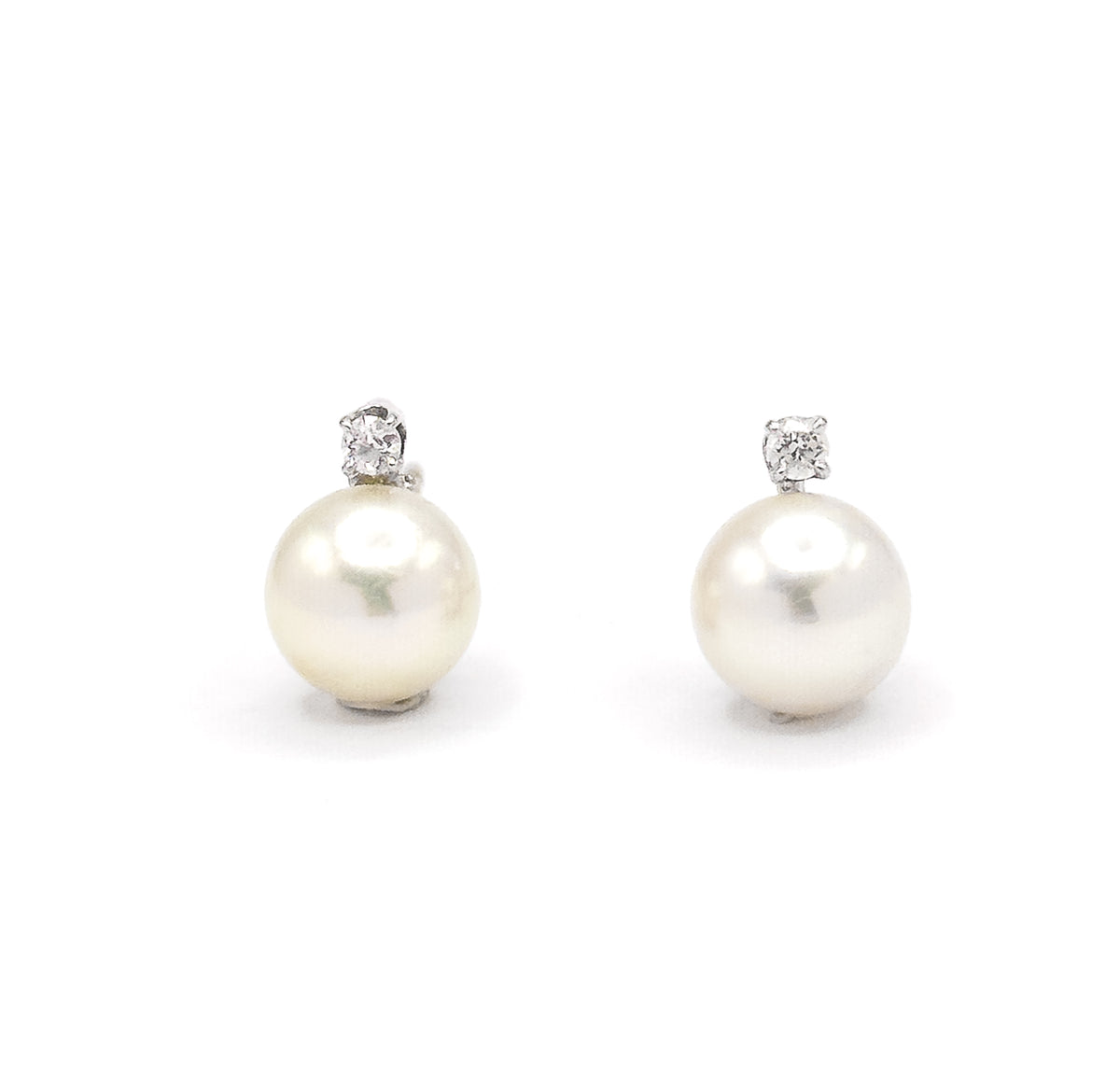 Both earrings face straight on side by side against white, each round creamy pearl topped by a prong-set diamond in white gold, matched in size and spacing.